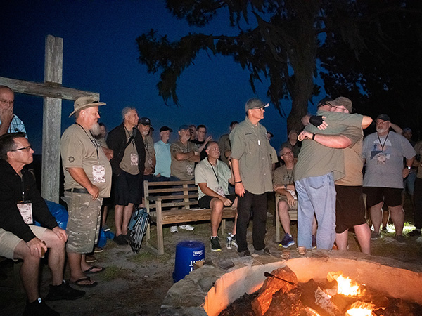 Men are standing around the bonfire at the Men's Boot Camp retreat. A large wooden cross is visible